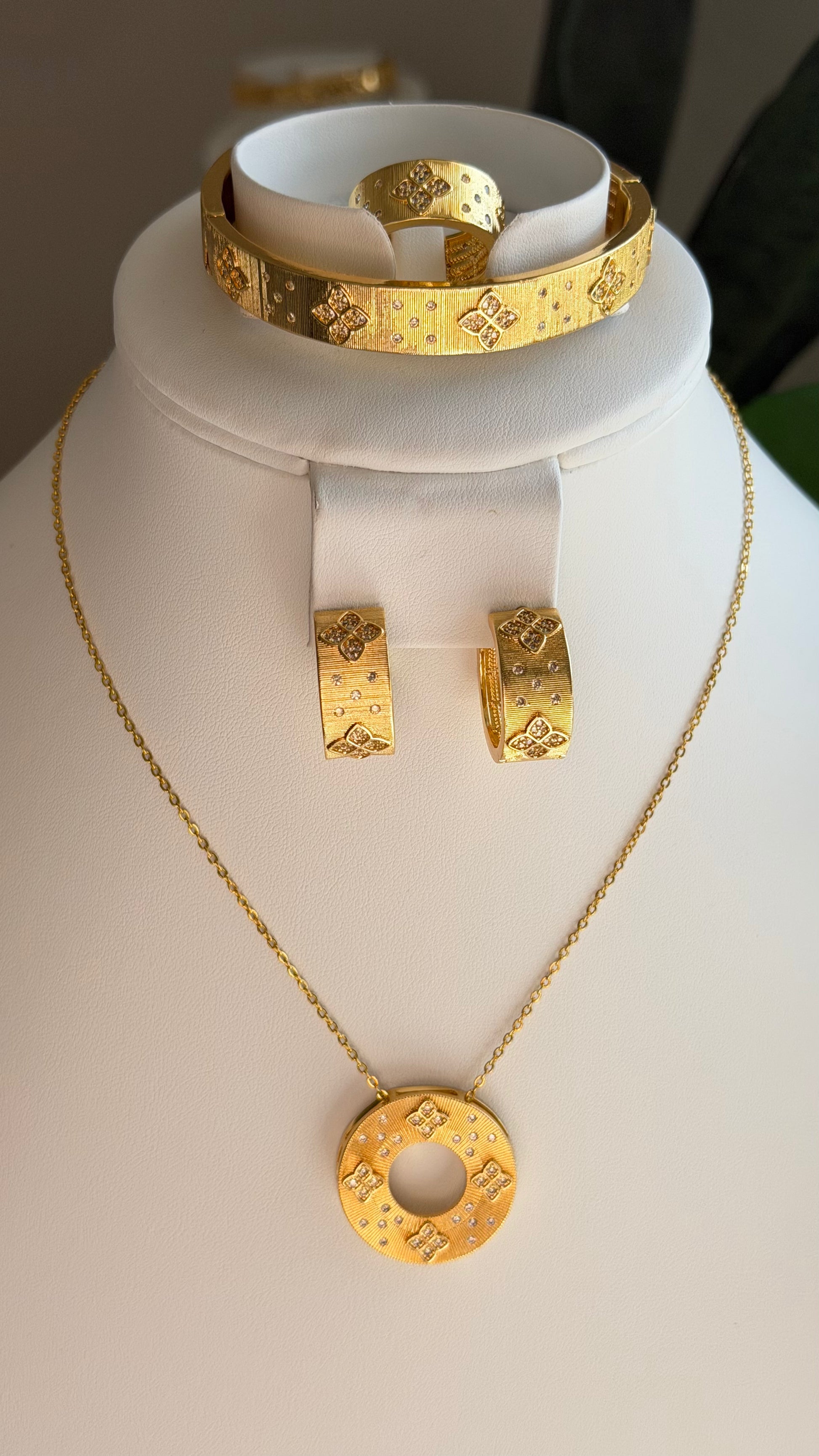 A gold jewelry set displayed on a white mannequin bust, including a necklace with a circular pendant, matching hoop earrings, and a cuff bracelet, all featuring an embossed monogram pattern.