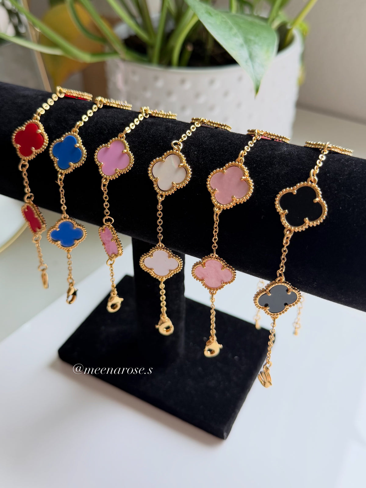 A set of gold-tone Alhambra-style four-leaf clover chain bracelets displayed on a black velvet jewelry stand. The bracelets feature colorful clover motifs in red, blue, pink, cream, and dark gray, with some pieces having dangling gold charms.