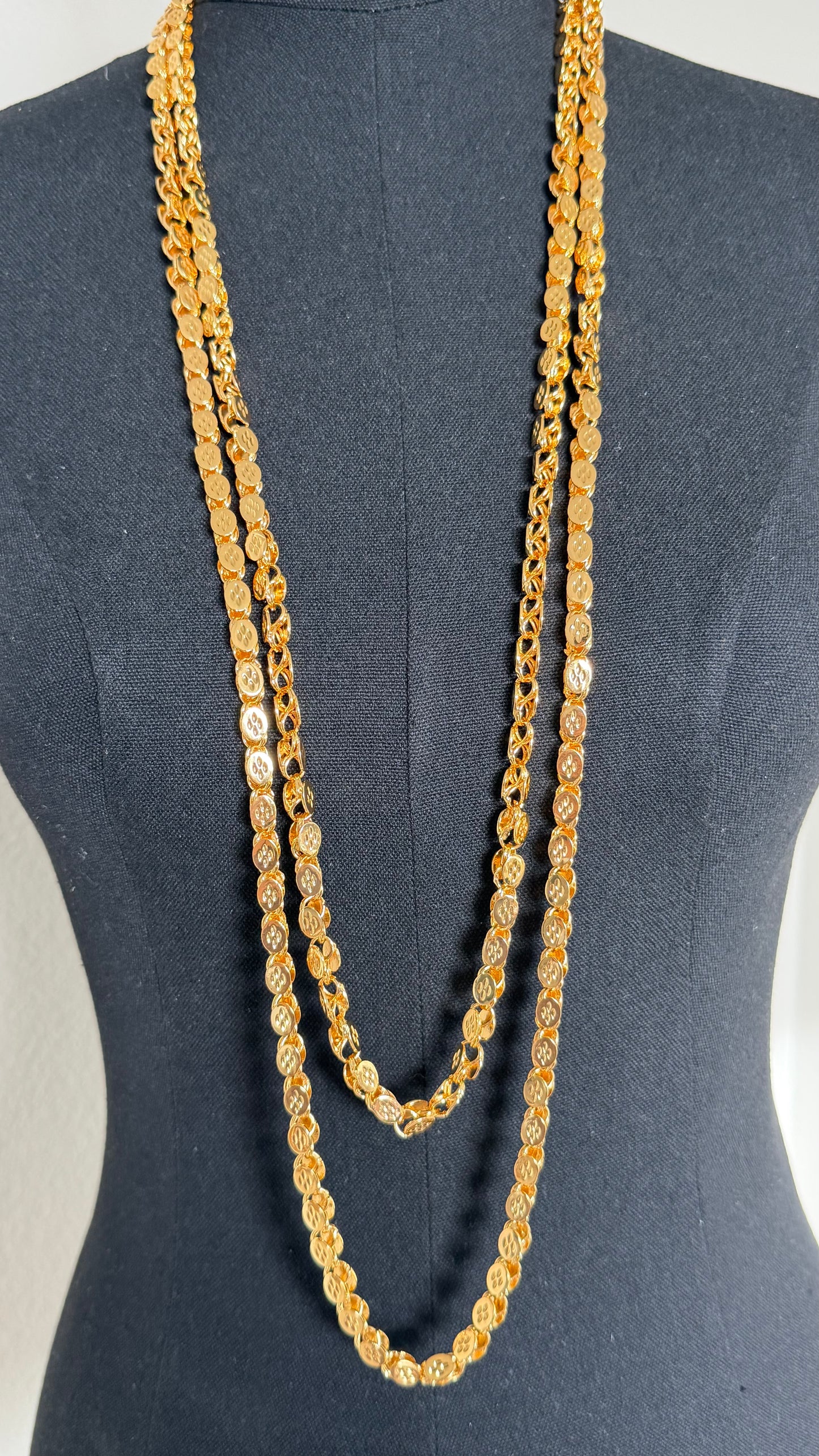 A long, layered gold chain necklace displayed on a dark gray mannequin torso. The necklace features three overlapping strands of textured, coin-like gold links.