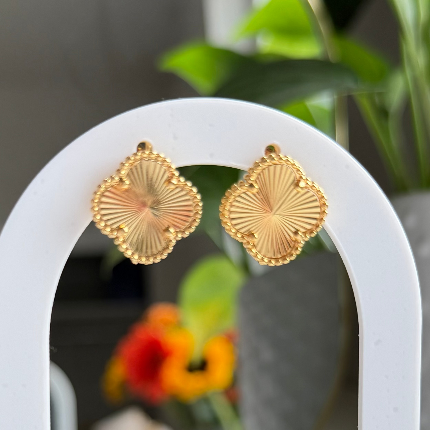 A pair of gold fan-shaped stud earrings displayed on a white arched stand with a wooden base, set against a softly blurred indoor background with green plants and warm decor.