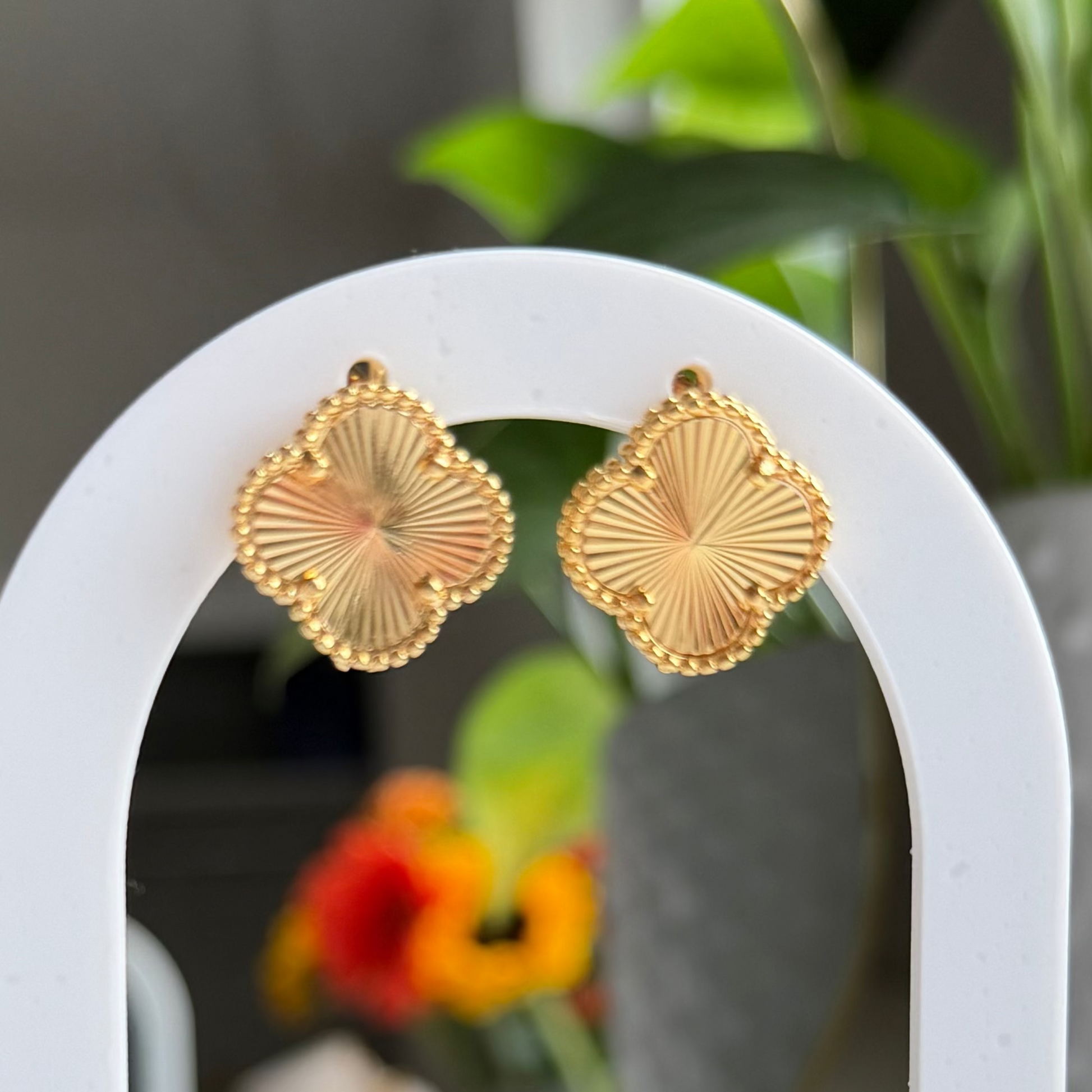 A pair of gold fan-shaped stud earrings displayed on a white arched stand with a wooden base, set against a softly blurred indoor background with green plants and warm decor.