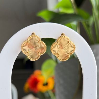 A pair of gold fan-shaped stud earrings displayed on a white arched stand with a wooden base, set against a softly blurred indoor background with green plants and warm decor.